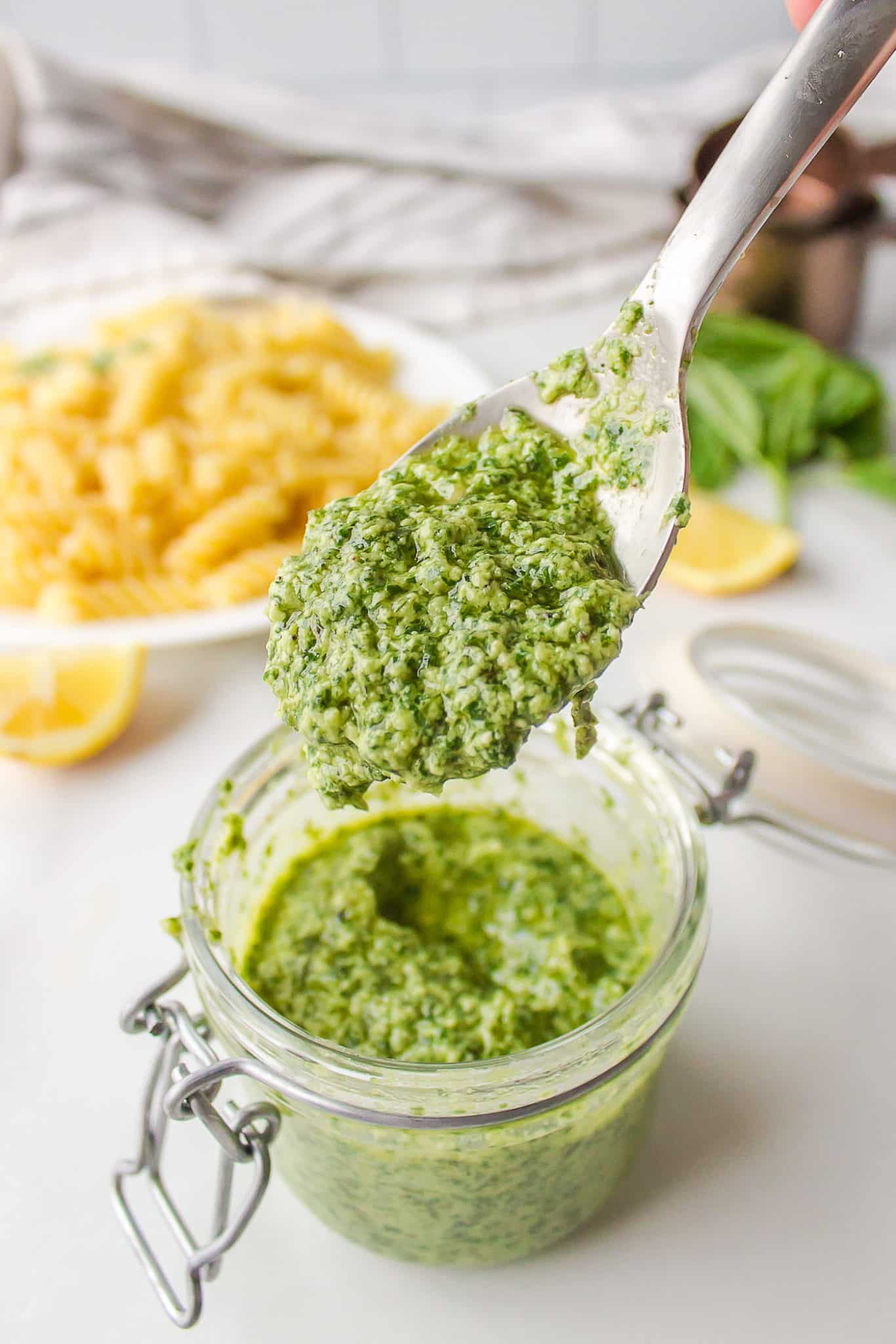 A spoon holding up a scoop of homemade pesto from a glass jar.