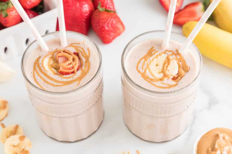 Two glasses of strawberry banana smoothie two straws and peanut butter drizzled on top.