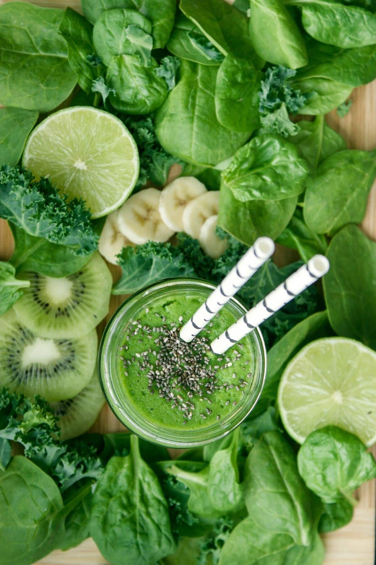 A green smoothie on a wood board surrounded by green fruits and veggies.