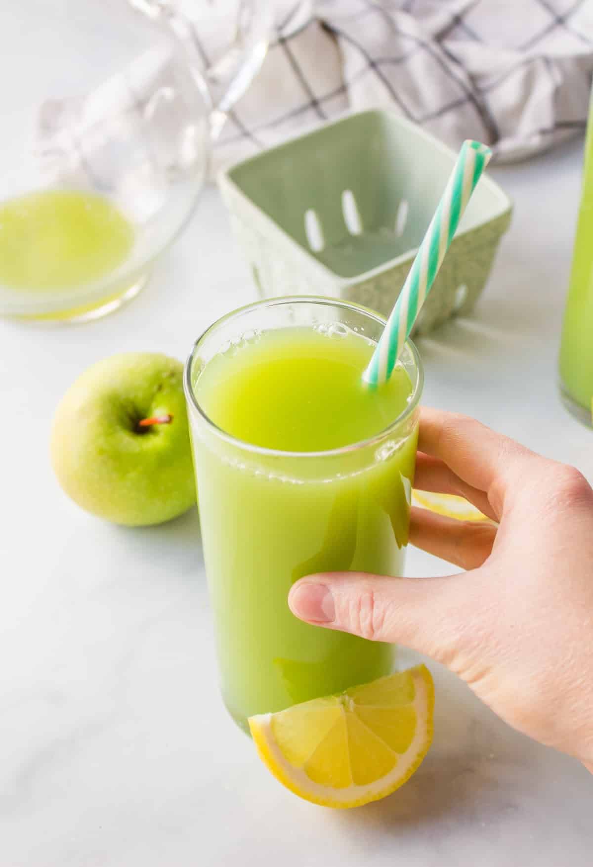 hand reaching for tall glass of pretty green juice with fresh lemon wedge.